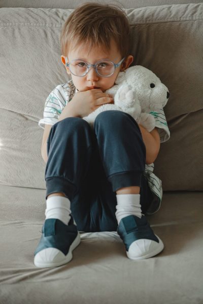 A child with autism in glasses sits on the sofa and is sad, hugs a soft toy teddy bear